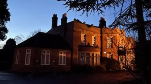 A two-storey building, pictured with light shining on part of the building that is protruding from the rest of the building on that side. A tree, which is dark on the image, is prominent in the foreground on the right.