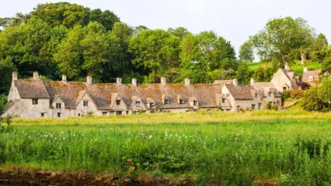 A famous line of small terraced Cotswold stone houses in Bibury, known as Arlington Row. The houses back onto a wooded area and, to the front of the properties, there is a large green space.