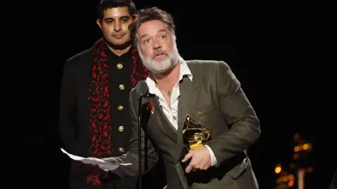 Getty Images Rufus Wainwright, wearing a grey suit and white shirt with open colour, holds a golden gramophone Grammy trophy as he holds a piece of paper and stands by a microphone. Behind him is a man dressed in a dark coat with a red and black scarf.
