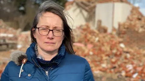 A woman in a blue puffy jacket looks at the camera. She has brown shoulder length hair and is wearing glasses. Behind her is a pile of red brick rubble and a partly demolished house.