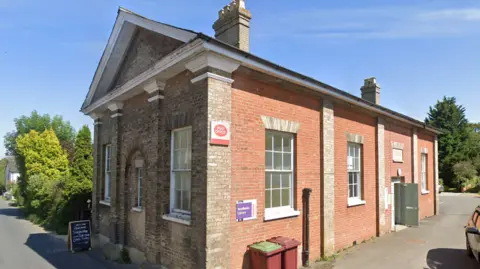 Google A general view of the exterior of a post office building. The single-storey building has a mixture of red and yellow brick. It has several large windows and a post office sign can be seen on its side.