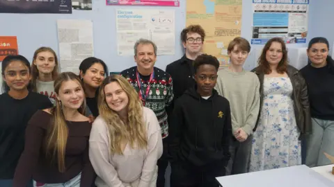A class of college students are standing in a group with their teacher smiling at the camera in front of a wall covered in science posters.