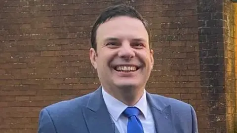 A man, with short brown hair, wearing a grey suit, white shirt and blue tie. He is smiling at the camera. There is a brown brick wall behind him. 
