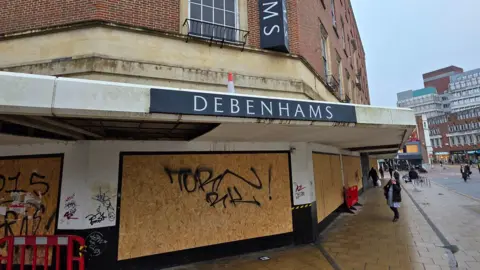 Paul Moseley/BBC One of the former entrances of Debenhams, now boarded up - with graffiti on the boards. A traffic cone has been placed on the roof covering the entrance.