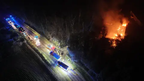 Eddie Mitchell A building covered in fire with its roof collapsed. The shadows of some trees can be made out around the fire. Multiple fire engines are lighting up a nearby road so yellow hoses can be seen.