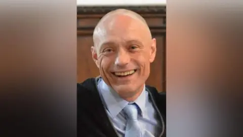 Family handout A bald and clean shaven man smiles at the camera. He is wearing a light blue shirt and tie and a black sweater