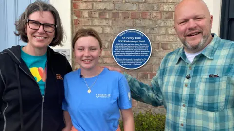 Jason Camus is a middle-aged man with a bald head and stubble, wearing a blue check shirt over a white t-shirt. He is standing alongside his daughter, Luciana, who is a teenager with brown hair tied back. She's wearing a gold necklace and a blue top and is standing next to her mother, Molly, who has a dark zip-up top and short dark hair. They are standing in front of a brick house and behind them a blue plaque says 55 Percy Park, WW2 Kindertransport Hostel. 