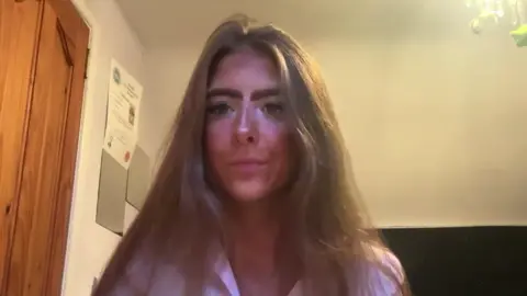 Louise has long brown hair and is wearing a pink striped shirt.