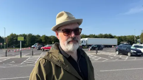 A man with a hat, sunglasses and coat standing at a car park.