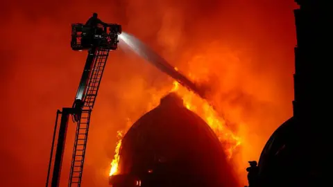 Getty Images A large domed building on fire. A firefighter on an extendable ladder is spraying water over the flames