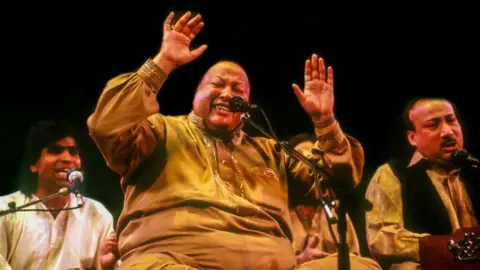 Getty Images A group of men performing. The main performer is sat cross legged and wearing gold-coloured trousers and a shirt with his hands raised in the air.