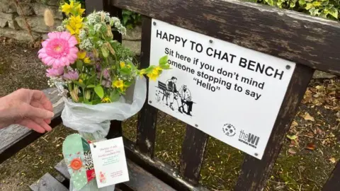 Anne Cradock Somebody placing a bunch of flowers on the corner of a wooden bench. The flowers are tied with a green patchwork cloth heart and note. On the bench is a sign which says it is a: "Happy to chat bench".