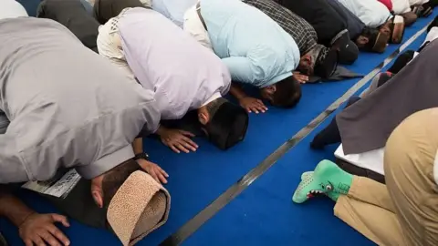 Getty Images Several people kneel in rows on a blue carpet during communal prayer inside a mosque, with heads bowed to the floor and prayer caps visible. The image is photographed from the side, showing multiple worshippers closely lined up during prayer.