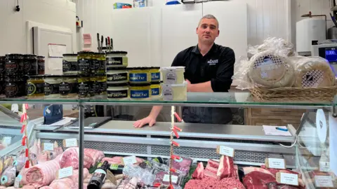 A man in a black shirt is standing behind a butcher's counter. 
