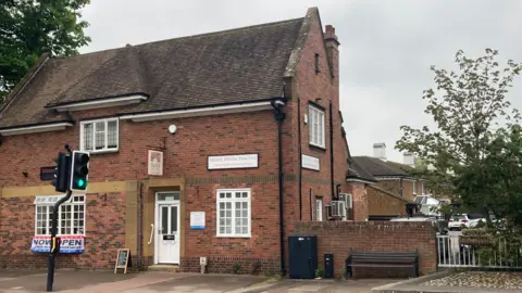 Laura Coffey/BBC A brick building with several signs on it. It has a white front door and three white windows on the front. There is a footpath running next to it and a traffic light in front of it.