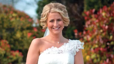 Family handout A woman in a white wedding dress smiles at the camera. She is holding a bouquet of white roses with both hands in front of her. Her blonde hair has been styled into curls, and a veil hangs from it behind her. The bride is pictured in a garden with a bench, medium level wall and bright bushes visible behind her.