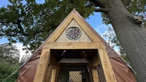 An image of The Gingerbread House, a structure built out of timber and plastic. It has a glass decoration at the top of the roof, which shows a brown heart with white and yellow dots around it  