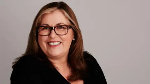 Mid-shot of Liza Tarbuck wearing a black top, leopard print frames and wearing streaks of blonde in her long flowing hair.