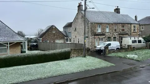 Carley Gilbert A residential street with a light sprinkling of snow settling on roofs and the grassy verges beside the pavement. 