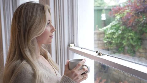 Sophie Richards looks out of a window holding a coffee mug