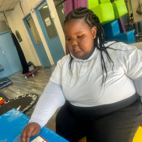 Memory Padi Sophia in school - she wears a white polo neck top and black trousers and her black hair is intricately braided. She sits at a table in a classroom and appears to be concentrating on a book in front of her