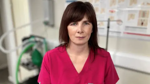 Dr Kate Hornby is wearing a dark pink veterinary uniform. She has long brown hair and is looking into the camera.