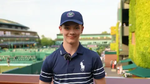 Alistair Durden / BBC A teenage boy wearing a blue polo shirt with white hoops on the bottom. He is stood in front of some tennis courts.
