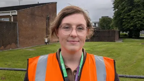 Dr Kerr has brown hair tied back, clear rounded glasses and is wearing a orange hi-vis with brown top and green lanyard.