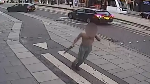 CCTV showing a topless man holding two large knives in one hand on a busy city street. His face has been blurred. There is a car parked haphazardly across a cycle land in the background.