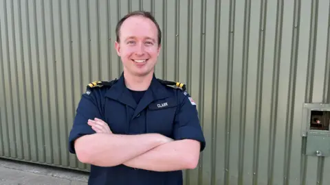 Lt Jack Clark stands in Royal Navy uniform against a corrugated iron background with his arms crossed and smiling directly into the camera