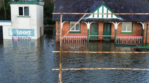 William Fisher The pavilion building is red brick and has green trimmings and door. The flood water is high along the front of the building.