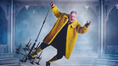 Patch Dolan Les Dennis swinging on a chandelier, wearing a yellow furry jacket with his mouth open inside the theatre