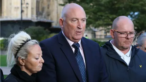 PA Media Bald-headed man flanked by woman with blonde hair and man with slight beard