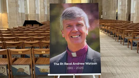 A photo of a bishop propped on a stand, captioned with "The Rt Revd Andrew Watson 1961-2026" at the bottom. It is placed inside a cathedral building lined with seats.