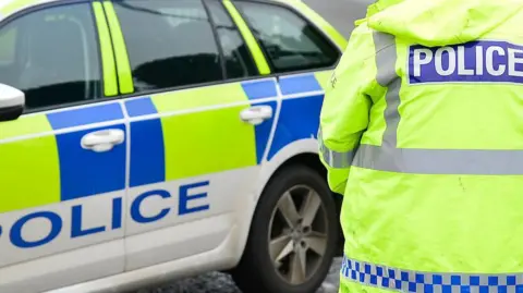 The picture is a generic one showing the back of a police officer wearing a fluorescent yellow jacket and standing beside a white, yellow and blue car
