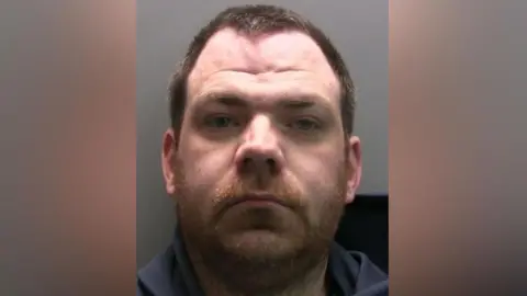Darrel Carter stares into the camera. He is a ginger coloured beard, short brown hair and has a widow's peak. He is wearing a navy hoodie.