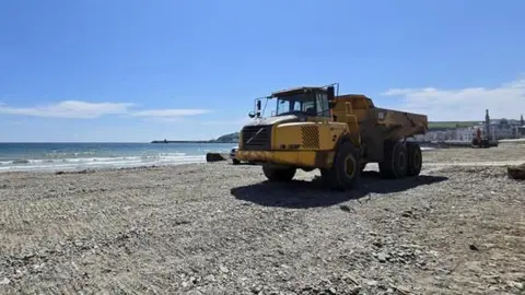A large yellow truck is parked on Douglas Beach. The top layer of sand has been removed.