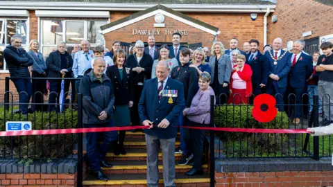 Telford & Wrekin Council A man wearing a suit with medals cuts a red ribbon in front of a brick building reading "Dawley House". There is a black fence either side of him with a red poppy sign to his right and a disabled wheelchair icon to his left. There is a crowd of people stood behind him.