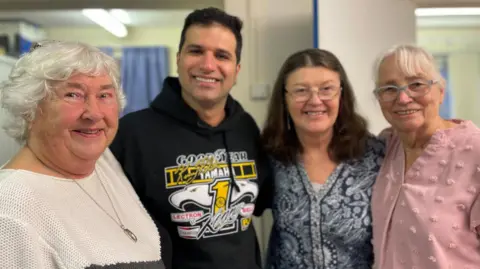 Simon Thake Three elderly women and one younger man wrap arms around each other smiling to the camera