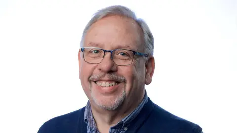 LDRS/Wakefield Council Pickersgill wears a dark blue sweater layered over a blue-and-white checkered button‑down shirt. He has short, light‑colored hair and stands against a bright, softly lit background.