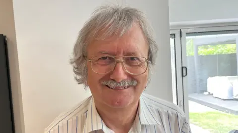 John Devine/BBC David Edwards has a full head of grey hair and matching moustache, he wears gold-rimmed glasses and a white stripped shirt. Behind him is a living room wall is a grey vertical radiator.