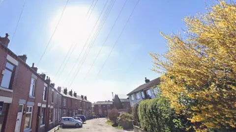 A streetview of Ormrod Street in Bolton. The road surface is damaged with potholes and looks like it has not been repaired regularly. On the left hand side is a row of two-storey red brick terraced houses which curve inwards slightly at the far end. They open directly on to a narrow pavement and cars are parked at right angles (so facing into the street) outside. On the right a semi-detached pair of more modern two-stoey houses can be seen peaking up behind hedges. At the end of the road there are more of the terrace buildings. It is a bright day with a hazy sun burning through what little cloud sits in the blue sky. 