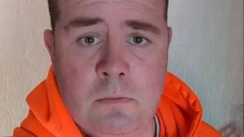 Dan Clarke has short brown hair and is clean shaven. He is wearing an orange hoodie.