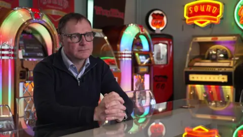 Image shows a man wearing a black jumper with a blue shirt underneath. He is sat at a counter with colourful juke boxes lit up behind him. 