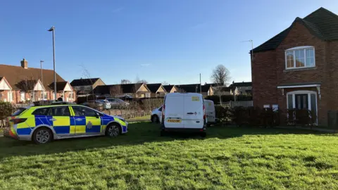 BBC A police car and other vehicles sit on the lawn of a brick house. It is a bright and sunny day.