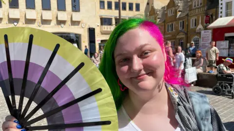 Sam Read/BBC Oskar Lee is standing outside with a fan green, white, purple and black fan held to the side of their face. They have medium length hair with a side part. One side is pink and the other is green. They are smiling at the camera while wearing pink earrings and a checkered black and white shirt.