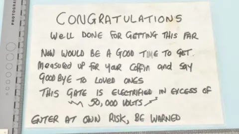 West Yorkshire Police A handwritten sign placed on a flat surface and framed by a photographic scale ruler. The sign is written in black marker on a piece of white paper, and the text adopts a dramatic, warning-style tone. It says:
“CONGRATULATIONS
Well done for getting this far
Now would be a good time to get measured up for your coffin and say goodbye to loved ones
This gate is electrified in excess of 50,000 volts
Enter at own risk, be warned"