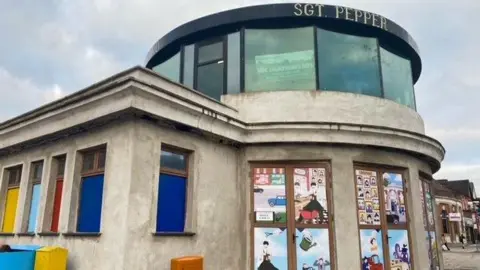 The Sgt Pepper's Bistro building is a partly round concrete structure with large glass windows running around the first floor. Artist Susannah-Rachel Gorman's Penny Lane-inspired artwork is contained within window panels on the ground floor.