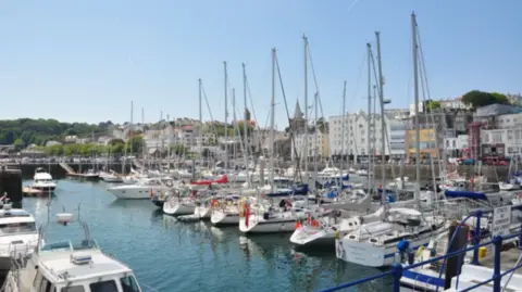 BBC A marina with yachts and other boats on one pontoon and other boats on another pontoon with houses behind and some trees in the background.