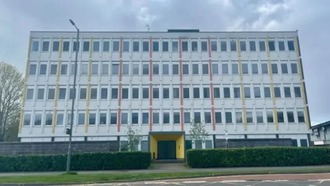 A five-storey block of flats with white, yellow and orange panels, and a yellow entrance, with a green hedge in front.